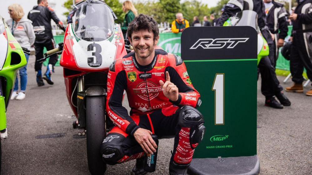 Mike Browne crouched on his haunches in front of his red race-winning bike holding up his left index finger in front of a green MGP sign with the number one on it in white. He has brown hair and is wearing black and red leathers.