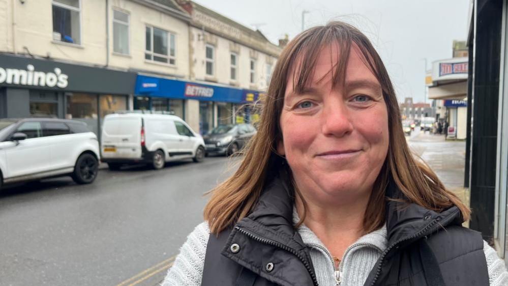 Emma Washbourne has long auburn hair and is wearing a grey jersey with a black padded gilet over the top. She is standing on Gorleston High Street, with parked cars and vans in the background, lined up in front of a parade of shops