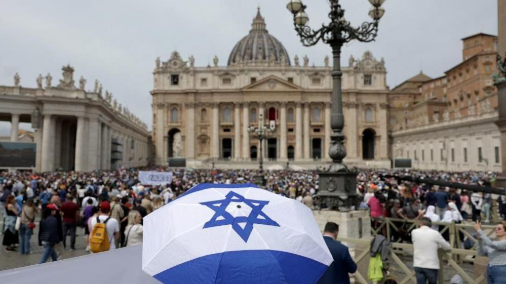 A person beneath an Israeli umbrella is seen standing near a large crowd 