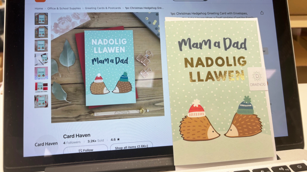 Nadolig Llawen Christmas card sold on Temu's website is on the left, and the similar card sold by Draenog is on the right. They both show hedgehogs nose to nose, wearing red and blue bobble hats, while snow falls. Temu's version reads "Nadolig Llawen Mam a Dad", while Draenog's reads "Mam a Dad Nadolig Llawen"