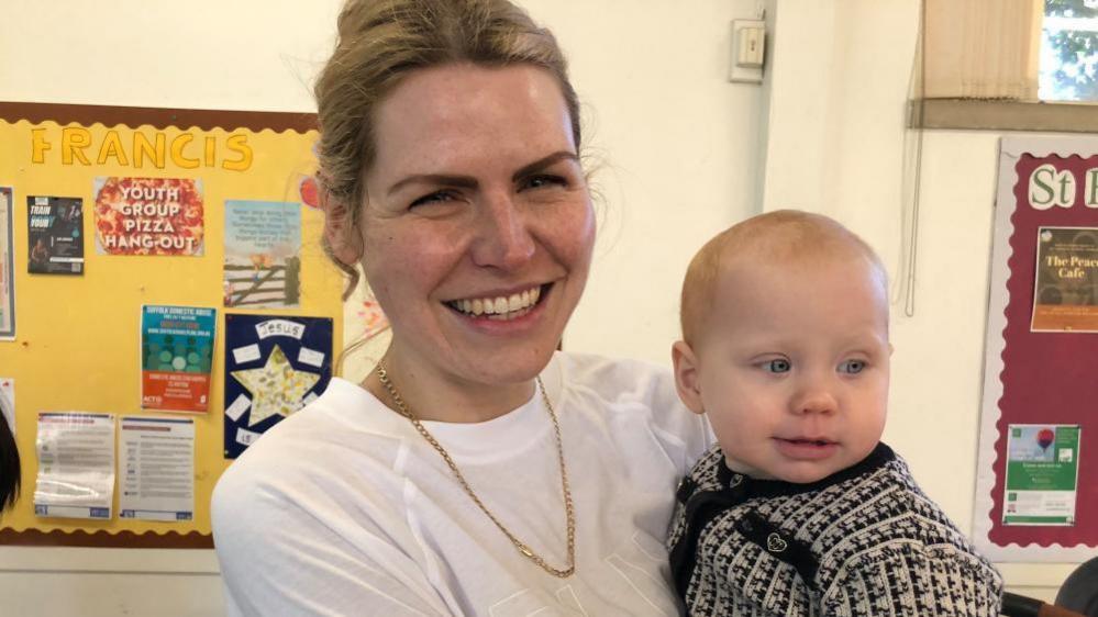 Martyna Green holding her Baby in her arms. She is smiling and wearing a white t-shirt. The baby has very blue eyes and is wearing a black and white patterned cardigan.  