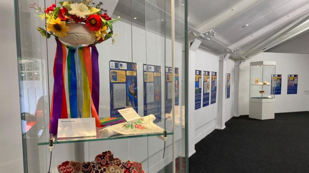 Image shows the inside of the exhibition - large prints of the artworks are displayed on the room's white walls. In the foreground a traditional vinok, a traditional Ukrainian flower wreath, is displayed in a case.