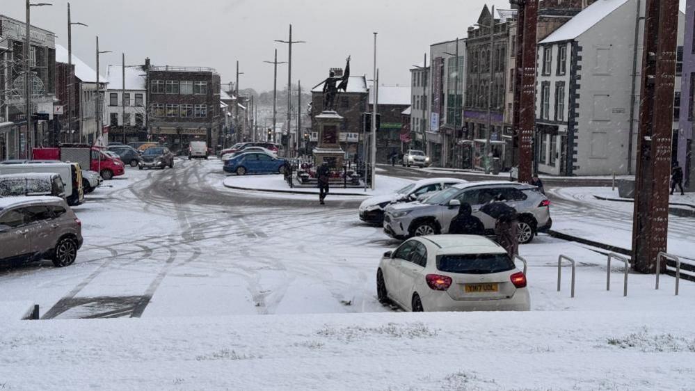 Dungannon town centre covered in snow. a number of cars can be seen parked up, while a man walks across the road