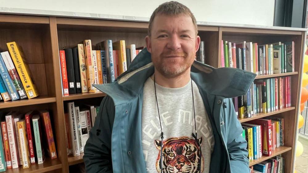 PJ Smith stands smiling in front of a bookcase at Spellow Library. He is wearing a blue coat and a grey sweatshirt with an image of a tiger. He has a beard and red hair.