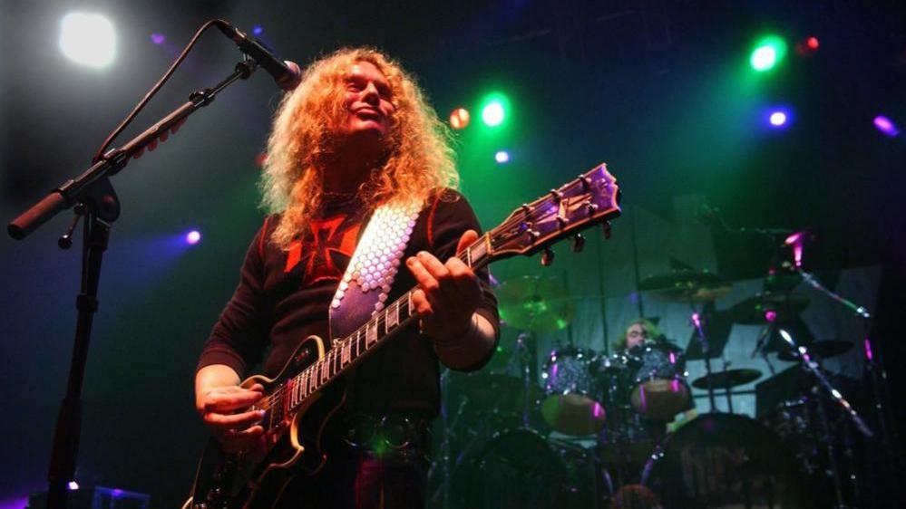 John Sykes playing with Thin Lizzy during a 2005 tour. He has long ginger hair and is playing a black Les Paul guitar supported by a wide silver strap. In the background a drumkit is visible. 