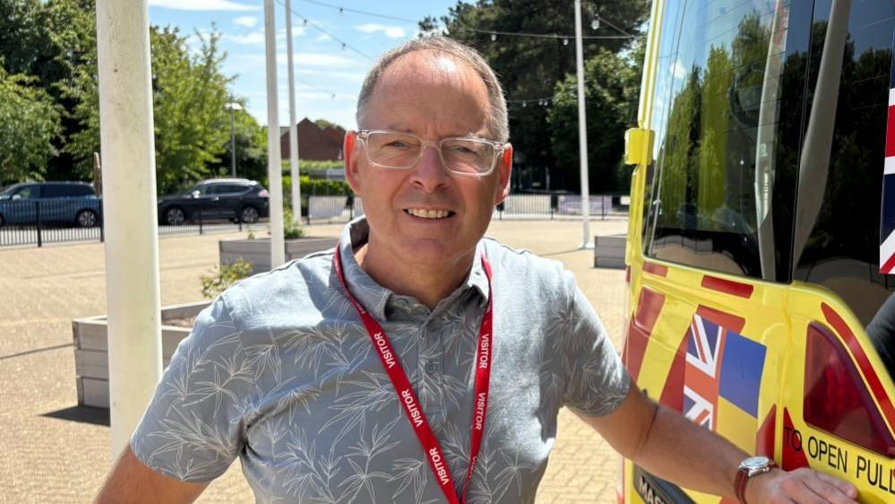 Steve Challen with short dark hair and glasses, looking at the camera and wearing a grey shirt and red lanyard. He is standing next to a yellow ambulance in the entrance to a school with several white pillars.