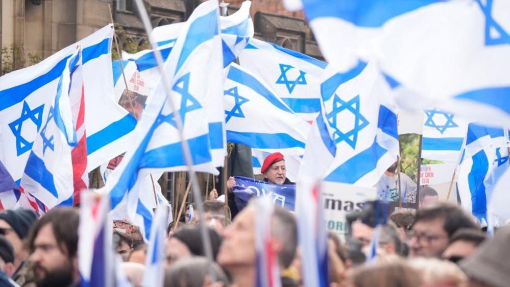 A large crowd of people gathered are waving white and blue Jewish flags. 