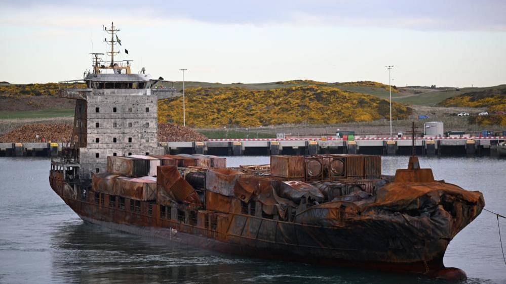 A container ship with most of its hull and its containers damaged by fire. In the background is a quay.