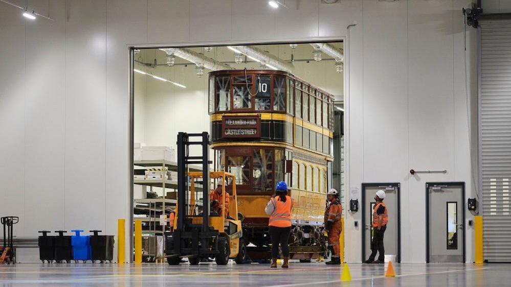 Inside a warehouse-type building, a large doorway with a forklift pulling through a yellow and brown vintage tramcar with two levels