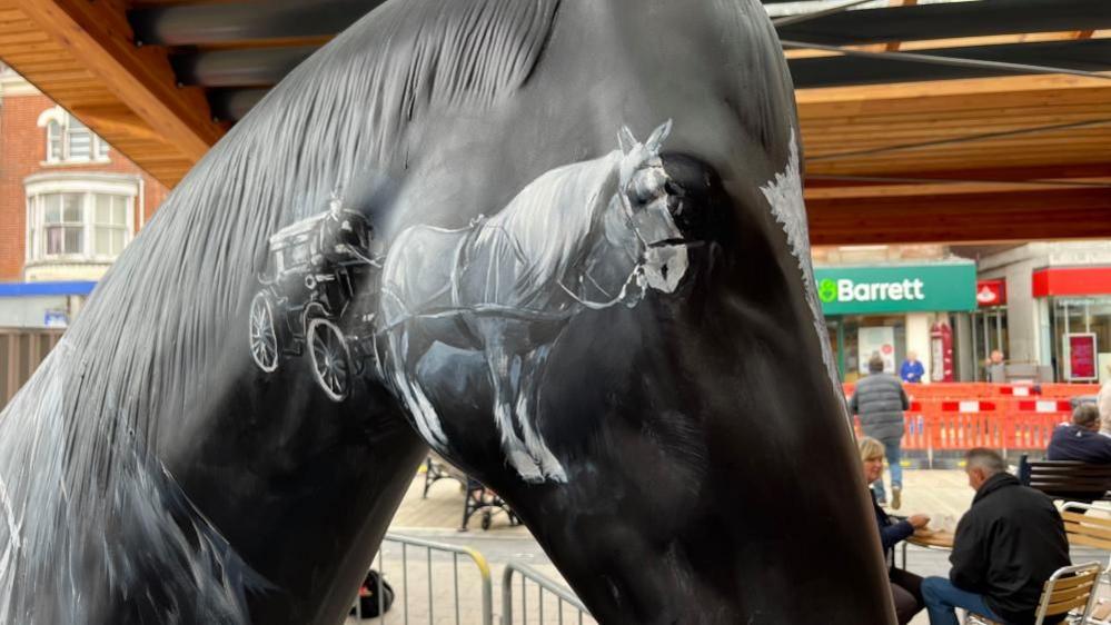 A horse's head, made of black fibre glass, featuring a depiction of a heavier horse pulling a coach and horseman. The picture is taken within Great Yarmouth Market, where the painting work was completed in front of the public during school holidays.