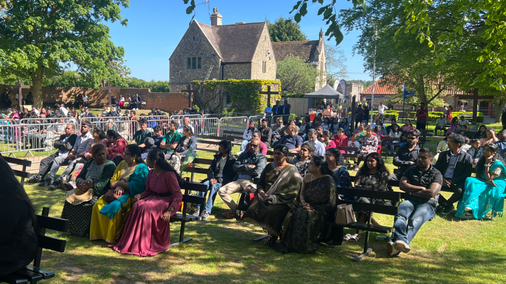 Lots of people are sat on benches in rows on a green field outside of the chapel. In the background is a stone house and two trees.