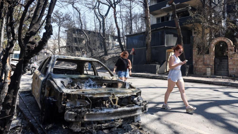 Women in shorts walk past a fully charred and burnt car ion a street surrounded by charred trees