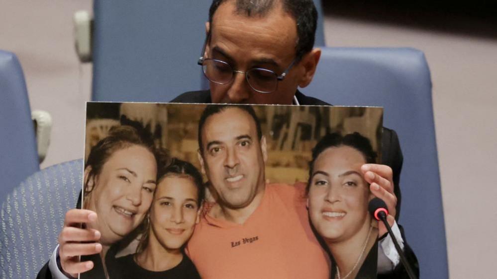 Eli Sharabi, his head downcast, holds an enlarged picture of himself with his wife and daughters.