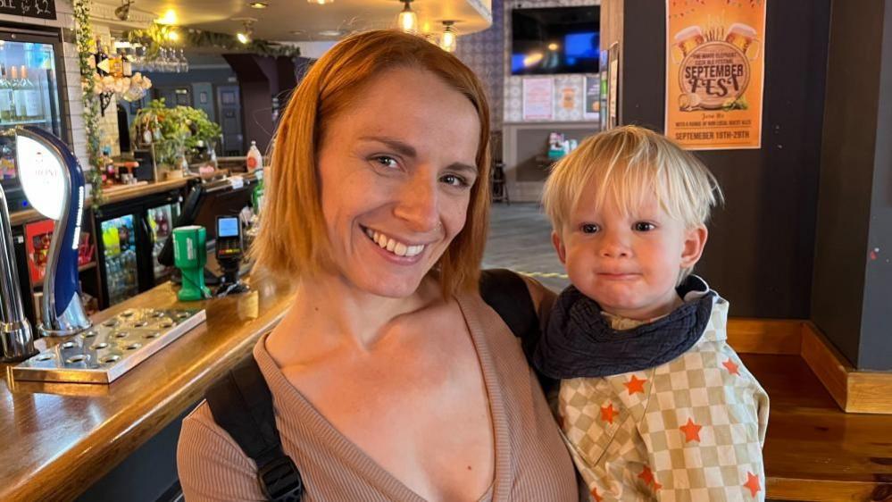 Lauren with medium-length brown hair, wearing a brown top and holding a child with blond hair wearing a green, white and red checked shirt in a pub.