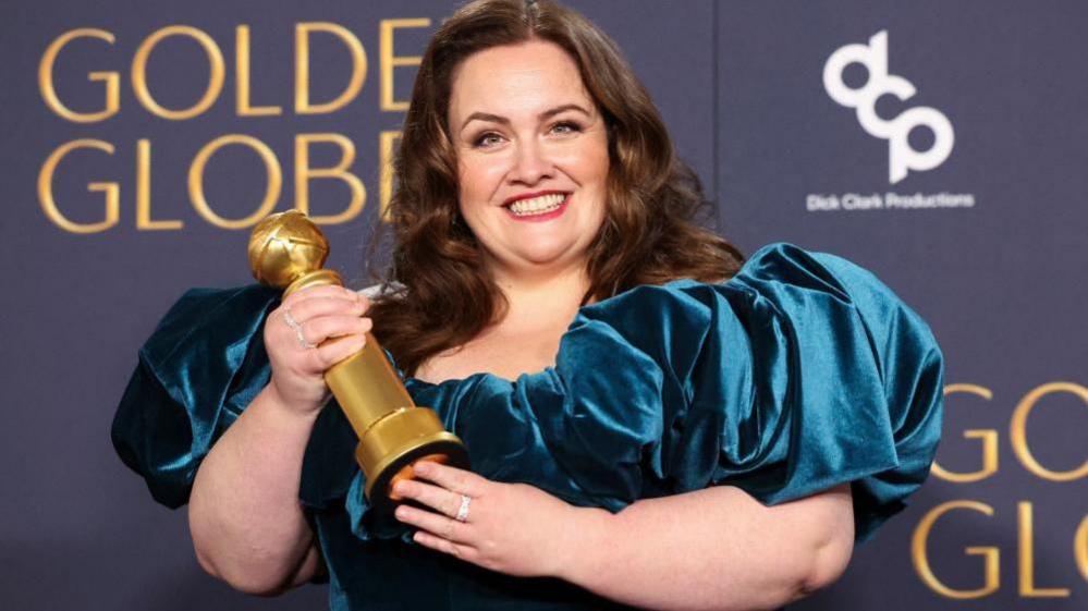 Jessica Dunning wins a Golden Globe award - she is seen with long brown hair and wearing a blue velvet gown, smiling and holding her award 