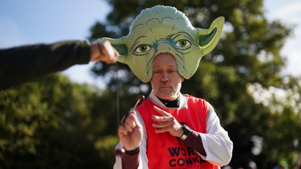 A man with a short grey beard and a large Yoda hat on his head striking an opponent's conker