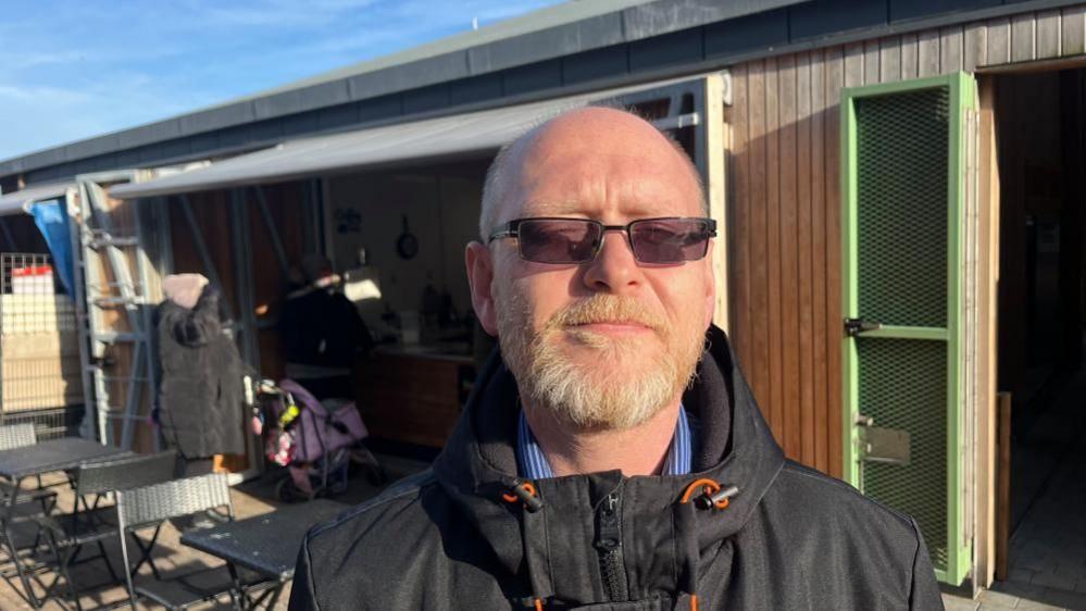 Paul O'Neill, wearing dark spectacles, has a whiting ginger beard and moustache. He is wearing a black jacket with orange laces around the hood, and a white-striped blue shirt beneath that. He is standing near the Great Yarmouth Market building; which is timber clad and has green doors.