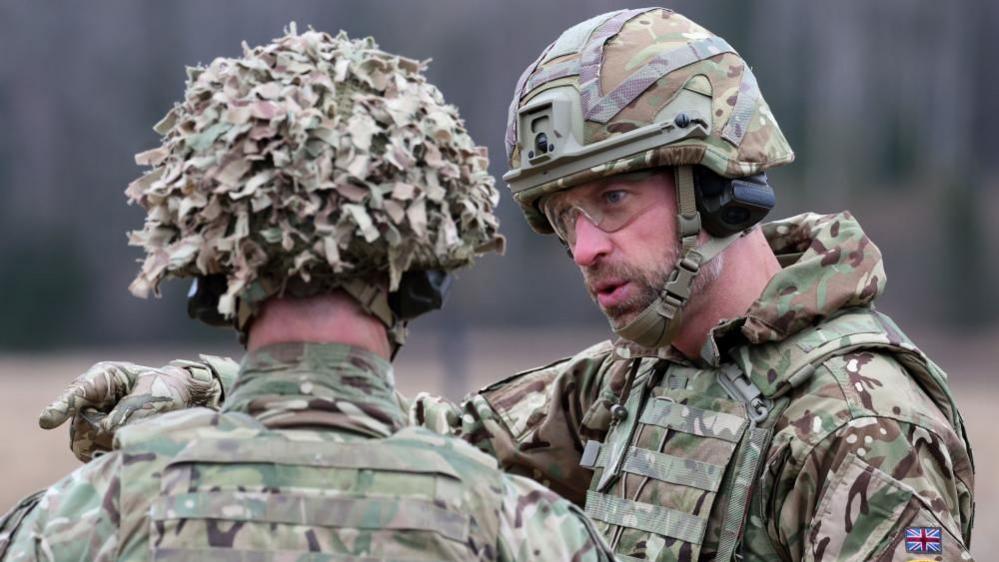 A man in military uniform, goggles and a helmet points while in conversation with troops.