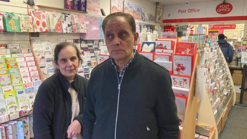 A postmistress wearing a black fleece next to the postmaster in a dark coloured top standing in front of greetings cards inside their post office.