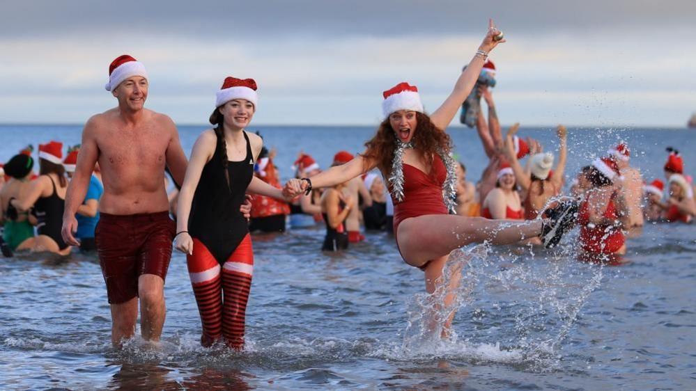 A man on the left is wearing red swim shorts and a red and white Santa hat. A girl in the middle has a dark coloured swimming costume on and is holding hands with a woman on the right who is kicking the sea water up. They are all smiling.