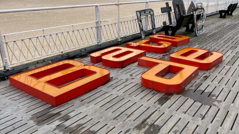 Letters B, a, t, a,  (upside-down) and e, made from orange acrylic, laid on the pier deck. The backing plates are stacked against a bench and railings. The beach is seen beyond the pier railings.