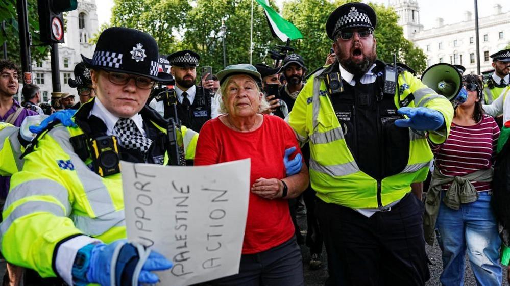 Two police officers escort a woman in a red shirt through a crowd