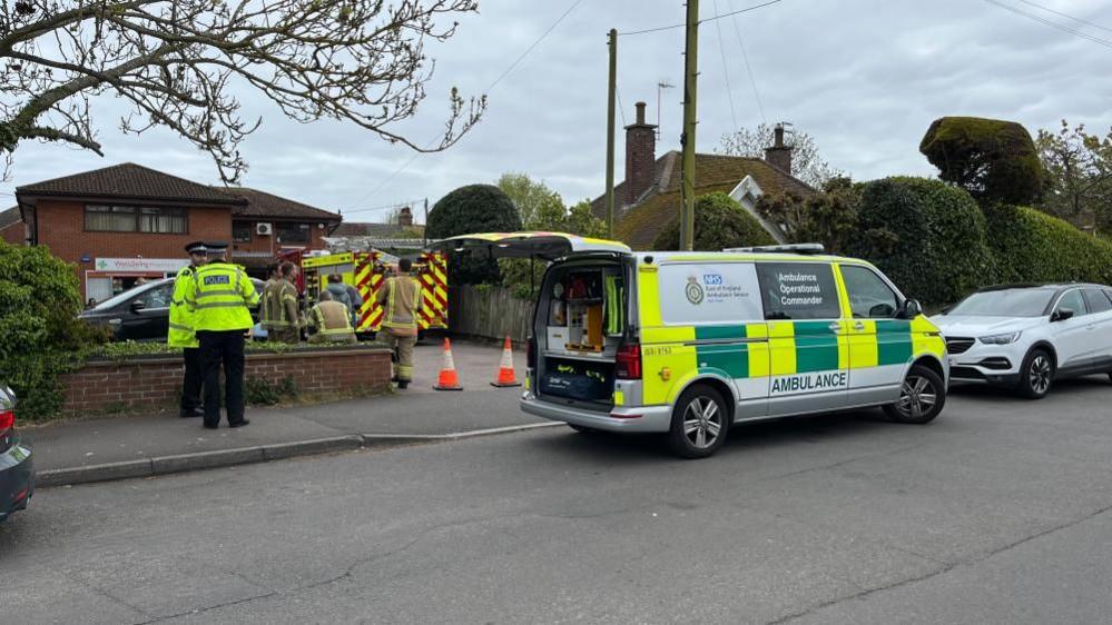 An ambulance parked outside Caister Health Centre, with the back door lifted open, and two police officers stand on the pavement. Two traffic cones are placed across the entrance to the medical centre car park, and a number of firefighters stand in the car park, with a fire engine on the car park outside the medical centre.