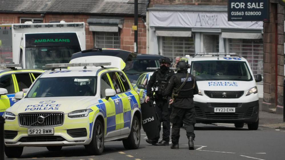 Two armed police officers in bullet-proof vests and wearing black helmets stand next to three police cars and a police van on the street
