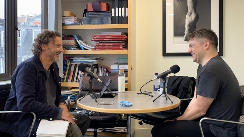 Julian Leeks (left) director of Bristol music charity Sound World speaks to a guest on his podcast series. They are sitting around a small round table that has two microphones on it. They are facing each other and both smiling.