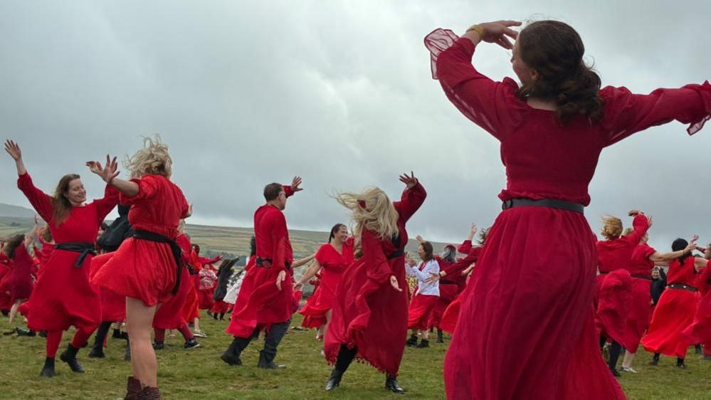 A group of people dressed in red robes dance in a grassy field.