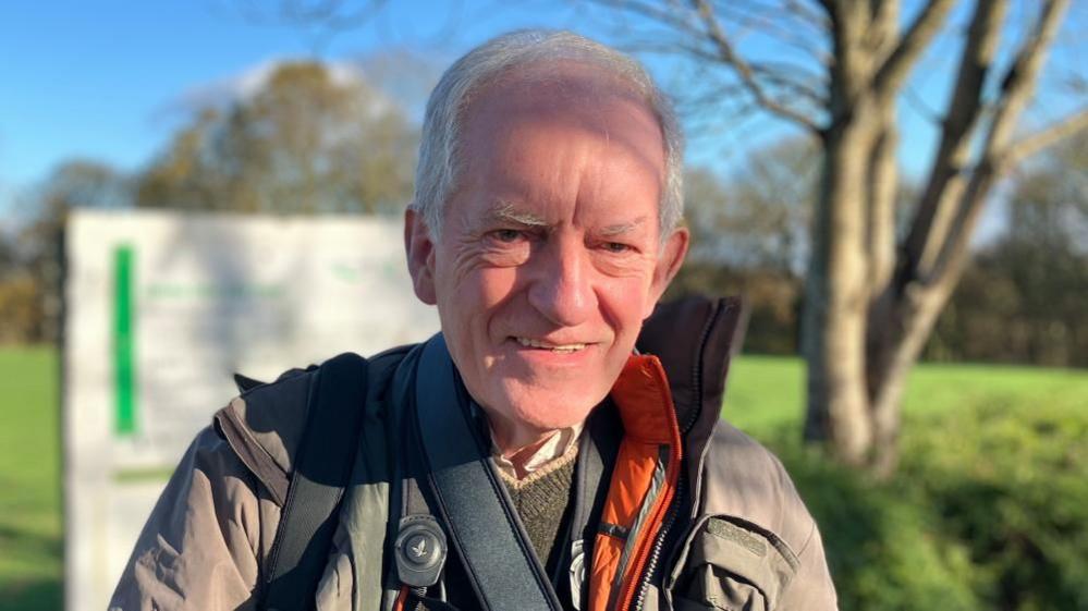 A man with short grey hair wearing a waterproof coat with birdwatching equipment over his shoulders. 