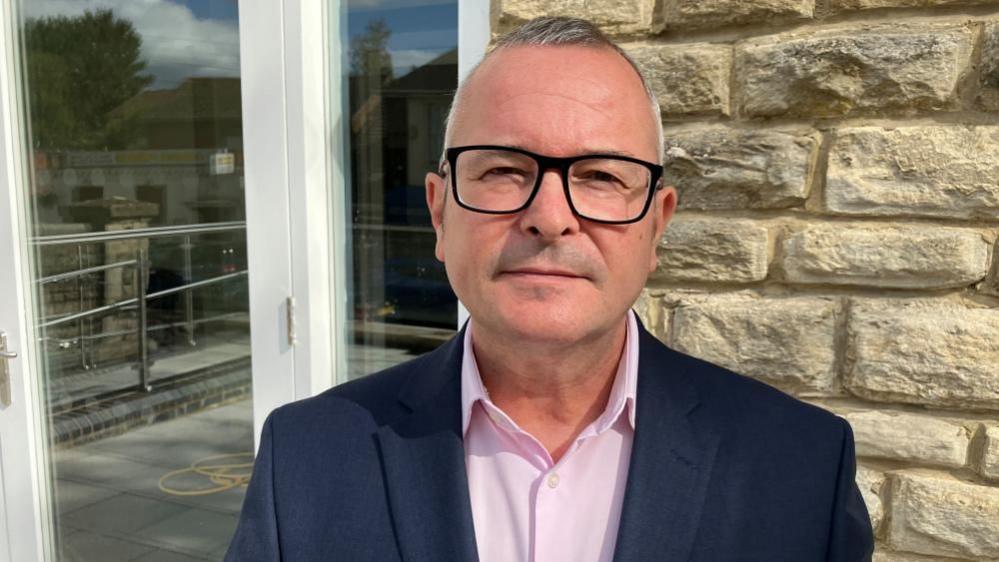 Lee Barron with short white hair and large black glasses looking at the camera wearing a dark blue jacket and pink shirt in front a stone wall with a patio-style door