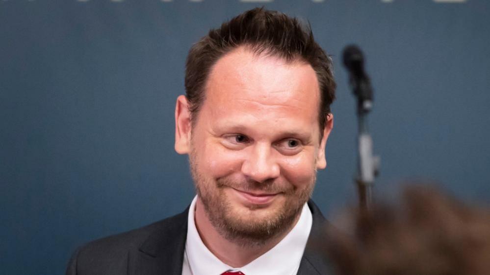 MP for Wakefield Simon Lightwood stands in front of a microphone, smiling at someone out of frame. 