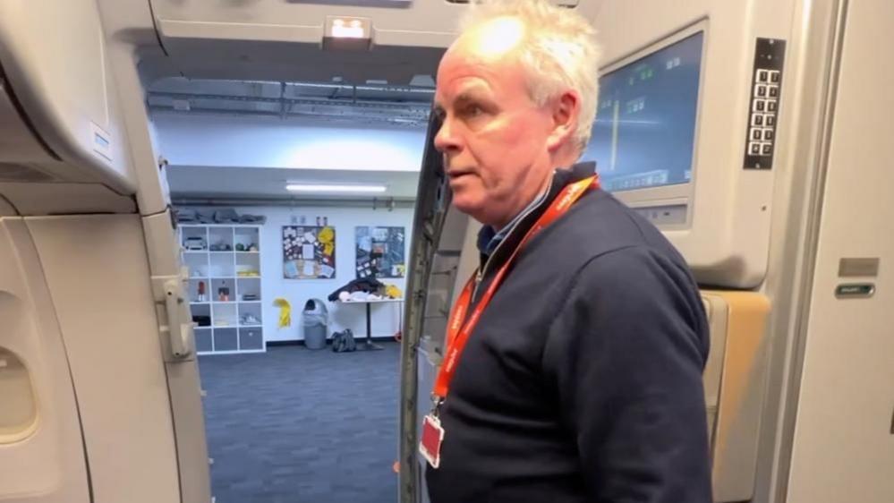 A 65- year-old man standing at the door of a training mock up of a plane. 