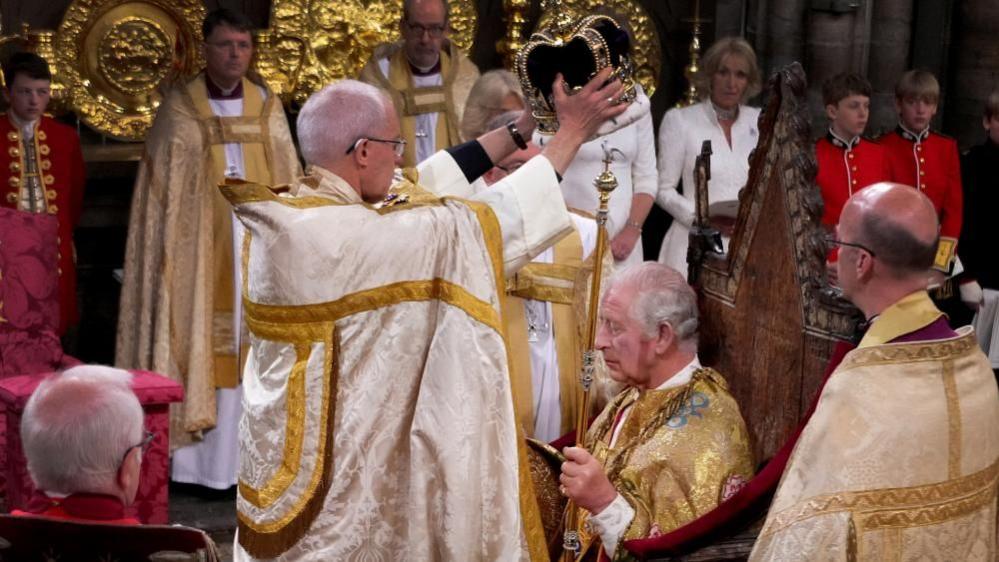 Archbishop Justin Welby holding a crown over King Charles's head, while Charles sits on a throne. Both are wearing golden robes.