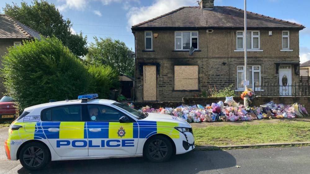 The house where the fire took place on Westbury Road, shown boarded up and with floral tributes outside. A police car is parked on the road in front.