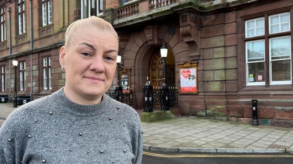 Emma Flaxman-Taylor has long blonde hair, tied back in a pony tail. She is wearing a grey jersey which has silver bobbles sewn on. She is standing outside Great Yarmouth Town Hall, a red-stone building dating to 1883 which features decorative masonry, sash windows and a large oak double door.