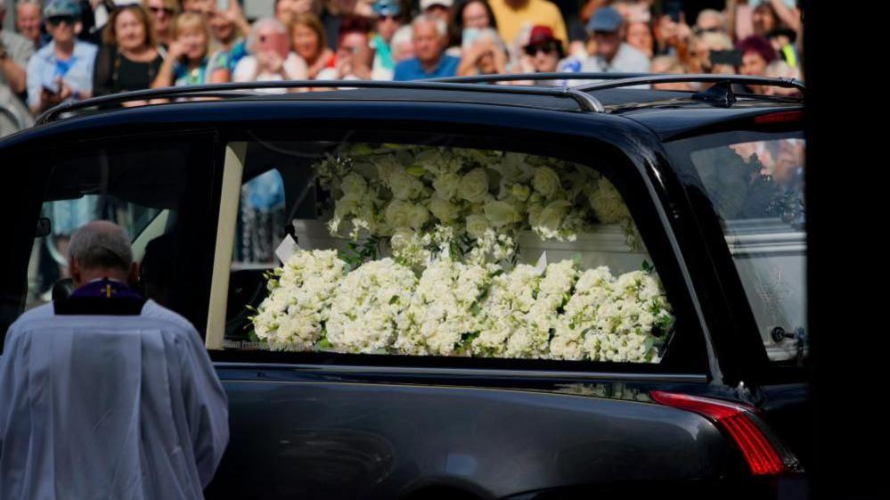 A floral tribute with white flowers spells out 'Bonnie'. White roses and lilies are placed on her coffin inside the hearse as it arrives at the church. A priest can be seen standing next to the hearse with his head bowed. Blurred out crowds of people can be seen behind the hearse.