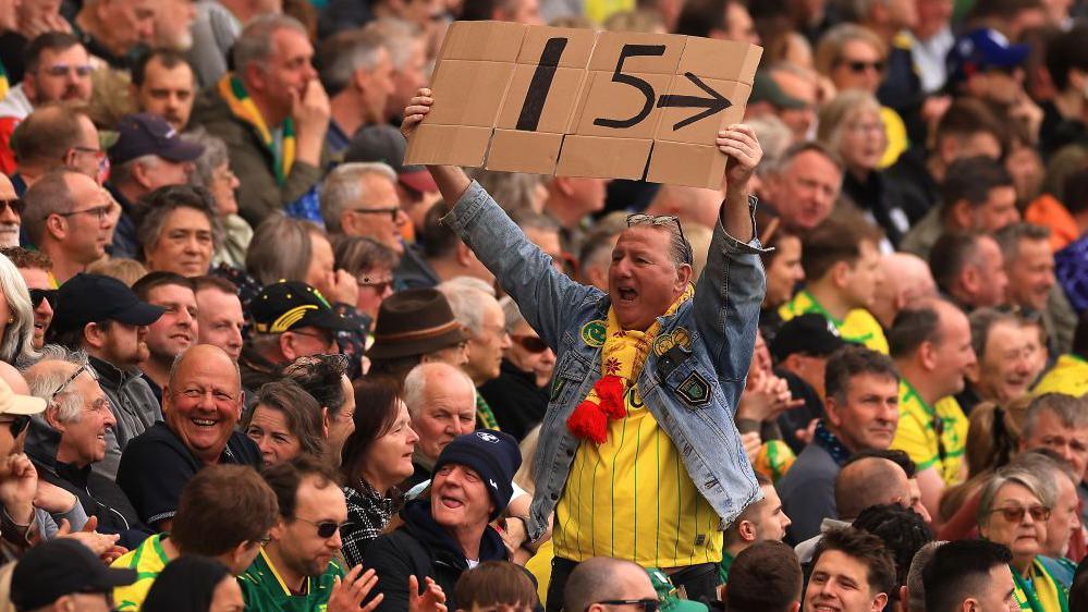 In a stand packed with fans who are seated, one fan is standing up. He is facing away from the pitch and is wearing a blue denim jacket over a yellow Norwich City shirt. He has his glasses on his head. He is cheering and holding a large cardboard sign over his head which reads "15", the number of years Norwich have gone undefeated in the derby.