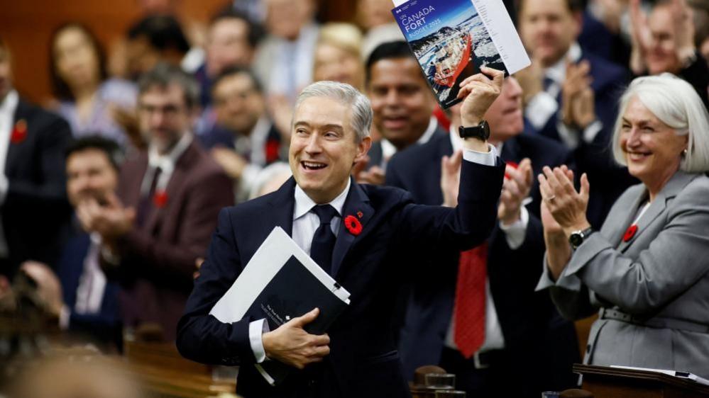 Canada's Finance Minister Francois-Philippe Champagne smiles as he holds up a copy of the federal budget in the House of Commons. He is wearing a dark suit and with a Remembrance Day poppy on his lapel, and other Liberal MPs applaud behind him