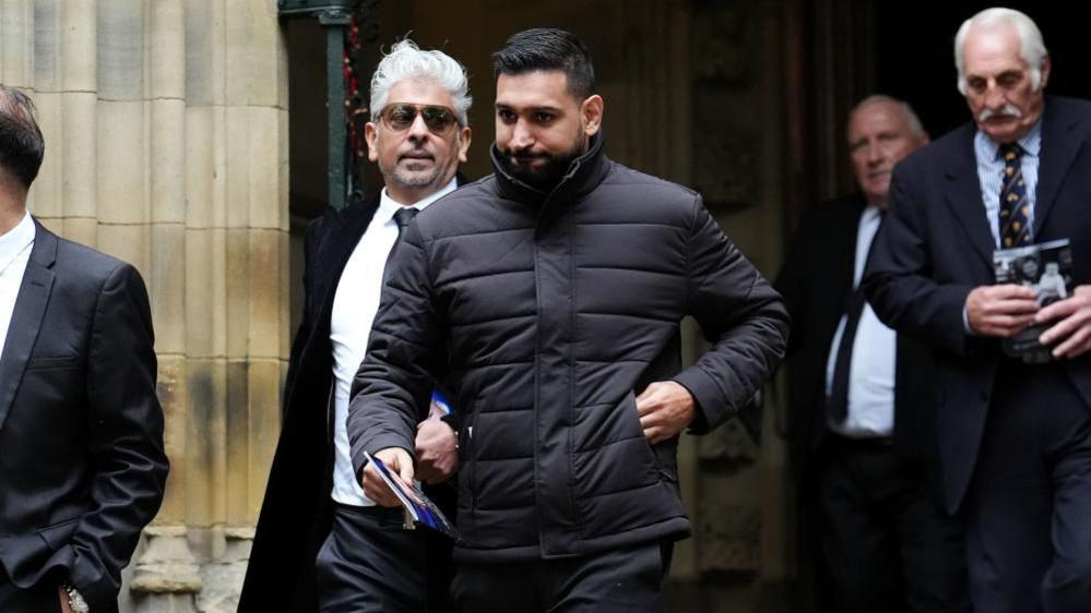 Amir Khan, wearing a black puffer jacket, hands a brochure in one hand and has his other hand in his jacket pocket as he walks out of a brick building.