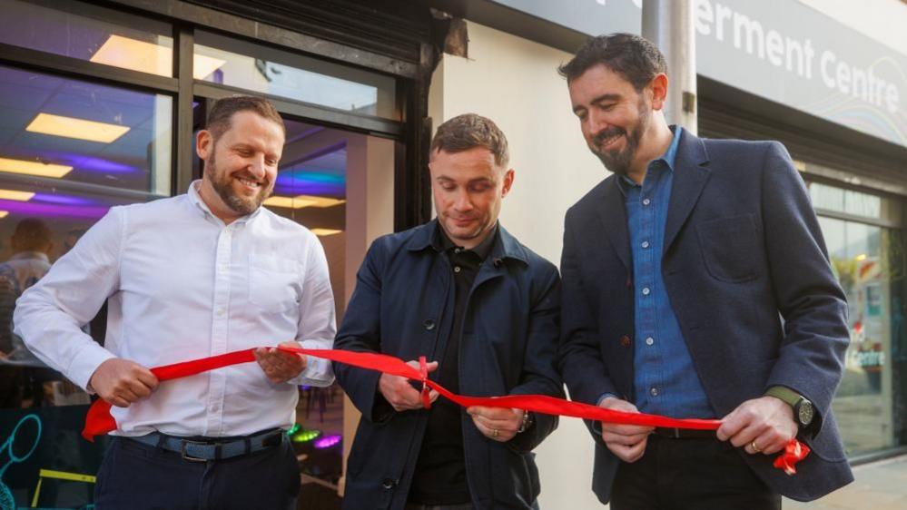 Three men are holding a red ribbon outside a building. The man in the middle is cutting the ribbon with scissors.