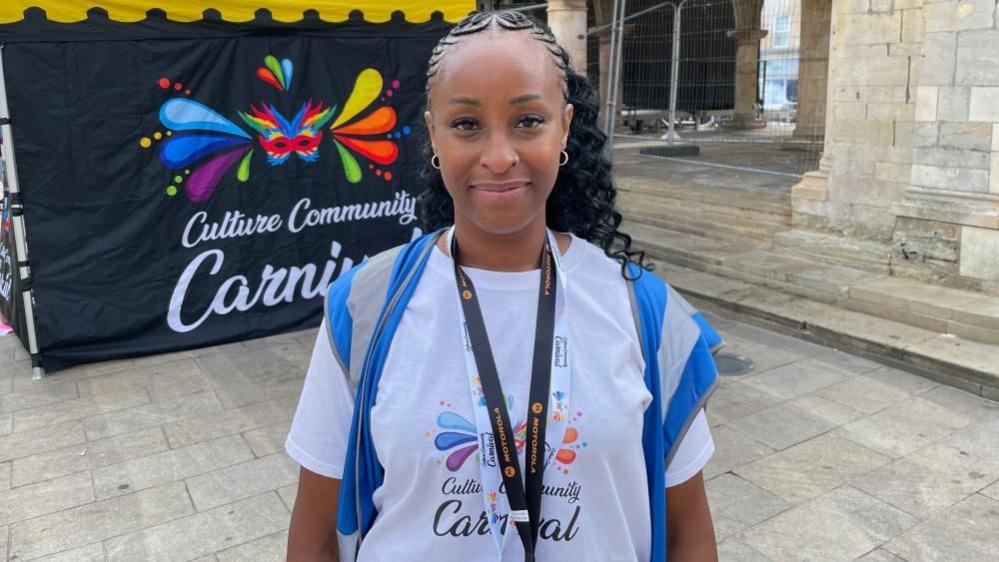 Precious Graham, standing in the centre of Peterborough, wearing a white T-shirt, she has a lanyard round her neck, she has long dark hair, has on a high-vis jacket, and is smiling and looking straight at the camera. A sign is behind her, and a brick building. 