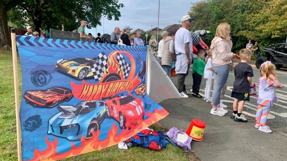 Adults and children stand alongside a road watching. Further back from the road is a flag with cars on it that says "Happy Birthday".
