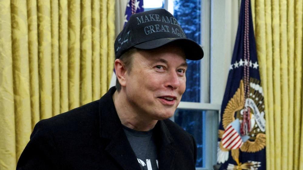 Elon Musk, wearing a black Make America Great Again hat, stands in the Oval Office.