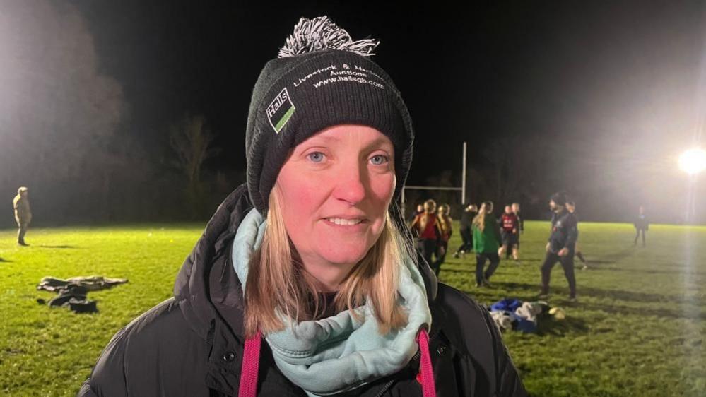 Sarah pictured looking into the camera and smiling slightly. She's stood on a flood-lit rugby pitch, which has green grass, a dark night sky, and is bordered by trees. She's wearing a black bobble hat, a black winter coat, and a light blue hoodie underneath. 