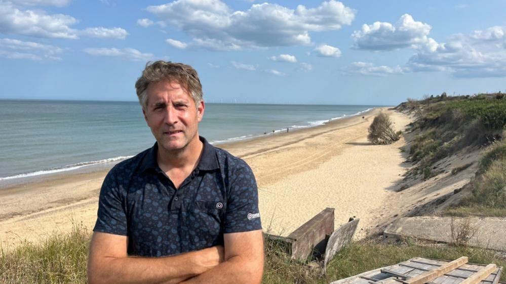 Simon Measures, standing with his arms folded across his chest, standing on the cliff in his garden at The Marrams in Hemsby. With medium length greying hair, he is wearing a patterned polo shirt, and behind him is Hemsby beach and parts of the cliff with debris left by erosion.