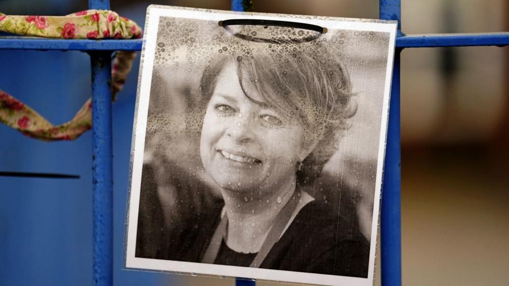 A laminated black and white photograph of a smiling Ruth Perry attached to a school railing. The photograph has water droplets on it. A child's headband is also attached to the railing.