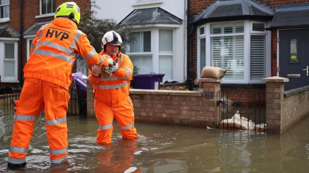 Wearing orange hi-vis coveralls two members of the Hertfordshire Fire and Rescue service create barriers with sandbags on a flooded street in Hitchen, Britain on 27th September 2024.