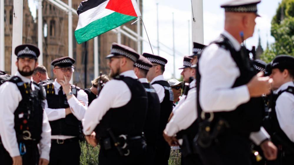 Police officers in white shirts, flat hats and stab-proof vests stand outside parliament during a protest, while a Palestinian flag is seen in the background